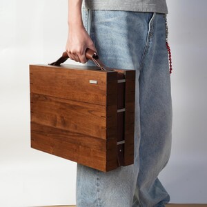 May include: A rectangular wooden briefcase with a smooth, dark brown finish, featuring a leather handle on top and a small silver-toned metal accent on the upper right corner. The case has visible joinery details along the side edges.