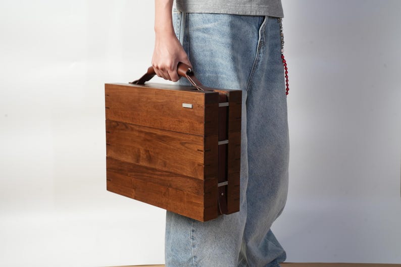 May include: A rectangular wooden briefcase with a smooth, dark brown finish, featuring a leather handle on top and a small silver-toned metal accent on the upper right corner. The case has visible joinery details along the side edges.
