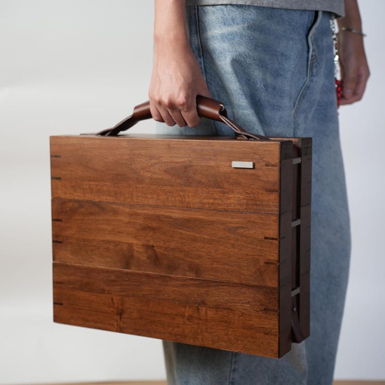 May include: A rectangular wooden carrying case with a dark brown finish and visible wood grain. The case features a brown leather handle attached to the top and a small, rectangular silver-toned metal accent piece on the upper right corner.