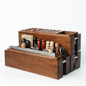 May include: A tiered wooden organizer for leatherworking tools featuring multiple circular slots and compartments. A metal ruler is positioned across the front, showing measurements in inches. The unit holds various brushes, wooden burnishers, and metal tools.