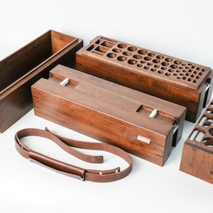 May include: A dark brown wooden storage box with a removable lid featuring numerous circular and square cutouts, a matching open rectangular box, a solid wooden lid, a separate wooden rack with circular holes, and a brown leather strap.