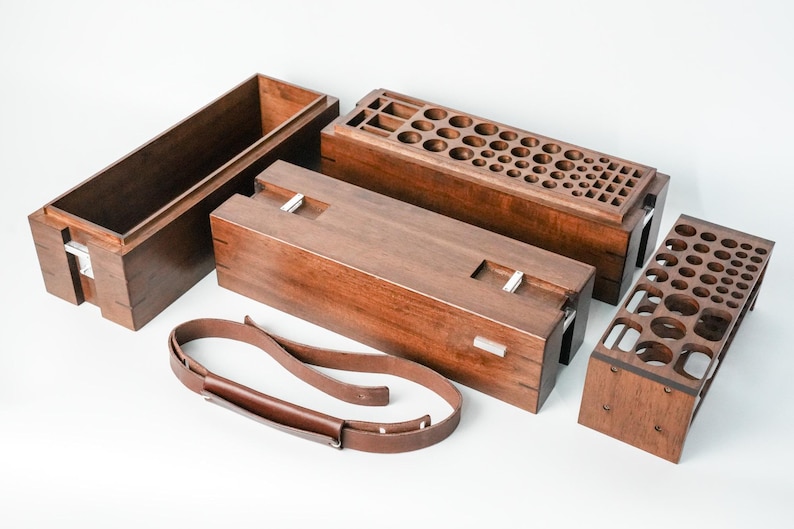 May include: A dark brown wooden storage box with a removable lid featuring numerous circular and square cutouts, a matching open rectangular box, a solid wooden lid, a separate wooden rack with circular holes, and a brown leather strap.