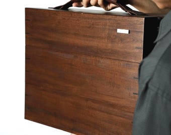May include: A rectangular dark brown wooden carrying case with a smooth finish and visible joinery details along the edges. The case features a dark brown leather handle attached to the top and a small, rectangular silver-toned metal accent piece.