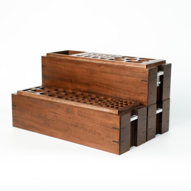 May include: A two-tiered dark brown wooden organizer featuring multiple circular cutouts on the top surfaces of both levels. The rectangular structure is constructed with visible joinery and includes small silver metal accents on the sides.