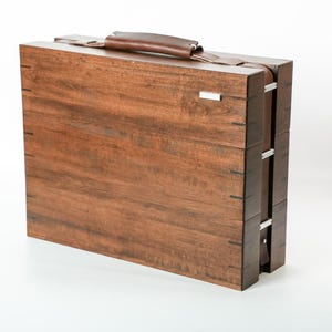 May include: A rectangular wooden briefcase with a dark brown wood grain finish, featuring a leather handle on top, silver-toned metal hinges along the side, and a small rectangular silver-toned metal accent piece on the upper right corner.