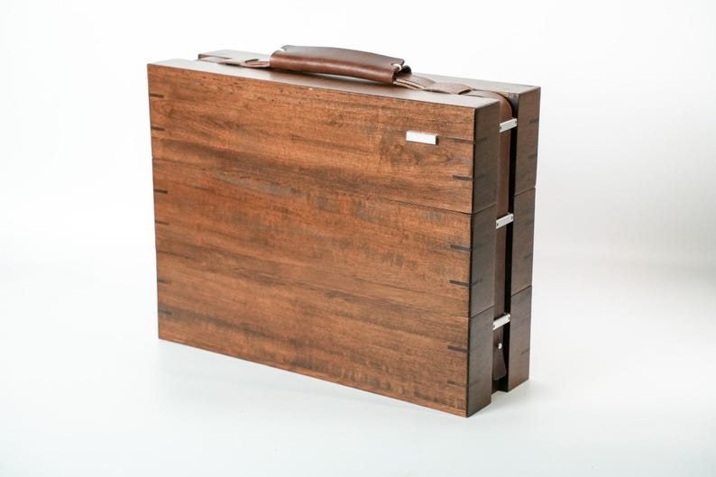 May include: A rectangular wooden briefcase with a dark brown wood grain finish, featuring a leather handle on top, silver-toned metal hinges along the side, and a small rectangular silver-toned metal accent piece on the upper right corner.