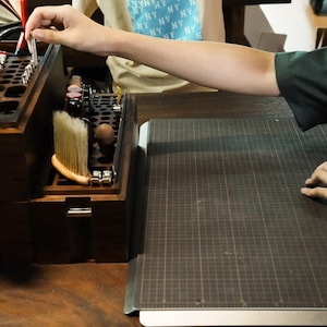 May include: A dark brown wooden craft organizer box featuring multiple circular slots for tools, a built-in brush, and metal hardware latches. The organizer is positioned next to a dark gray grid-patterned cutting mat on a wooden surface.