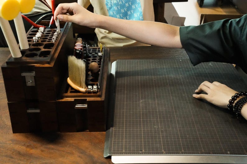 May include: A dark brown wooden craft organizer box featuring multiple circular slots for tools, a built-in brush, and metal hardware latches. The organizer is positioned next to a dark gray grid-patterned cutting mat on a wooden surface.