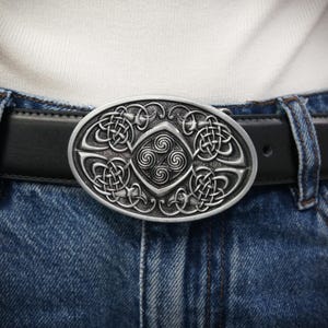 Könnte beinhalten: Ovale, silberfarbene Gürtelschnalle mit keltischem Knotenmuster. Die Schnalle ist an einem schwarzen Ledergürtel befestigt, der mit einer blauen Jeans und einem weißen Hemd getragen wird. Die Schnalle weist komplizierte Knotenarbeiten und ein zentrales Spiralmuster auf.