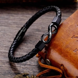 Puede incluir: Pulsera de cuero negro con un dije de símbolo de infinito grabado con los nombres "Tina" y "David".
