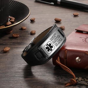 Op de afbeelding: Zwart leren medisch armband met een zilveren metalen tag gegraveerd met "Diabetes, Insuline, Allergieën, Penicilline, ICE: Tom G, 555-555-5555".
