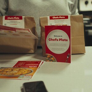 May include: A kitchen setting with two brown paper bags labelled "Chef's Plate", a red box with the same logo, and printed materials. The red box reads "Welcome to Chef's Plate". A person is holding a leaflet.