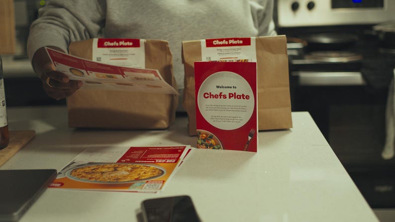 May include: A kitchen setting with two brown paper bags labelled "Chef's Plate", a red box with the same logo, and printed materials. The red box reads "Welcome to Chef's Plate". A person is holding a leaflet.