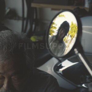 May include: A man with grey hair and a moustache is seen in a garage setting. A motorcycle mirror reflects a person with long hair and a white shirt. The scene is dimly lit, with a focus on the man and the motorcycle.