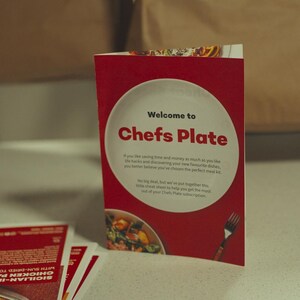 May include: A red and white Chefs Plate welcome card stands on a white surface, with the text "Welcome to Chefs Plate". The card features a plate graphic and a fork illustration. Other recipe cards are visible.