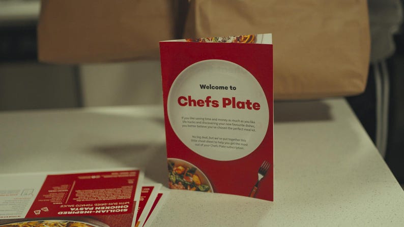 May include: A red and white Chefs Plate welcome card stands on a white surface, with the text "Welcome to Chefs Plate". The card features a plate graphic and a fork illustration. Other recipe cards are visible.