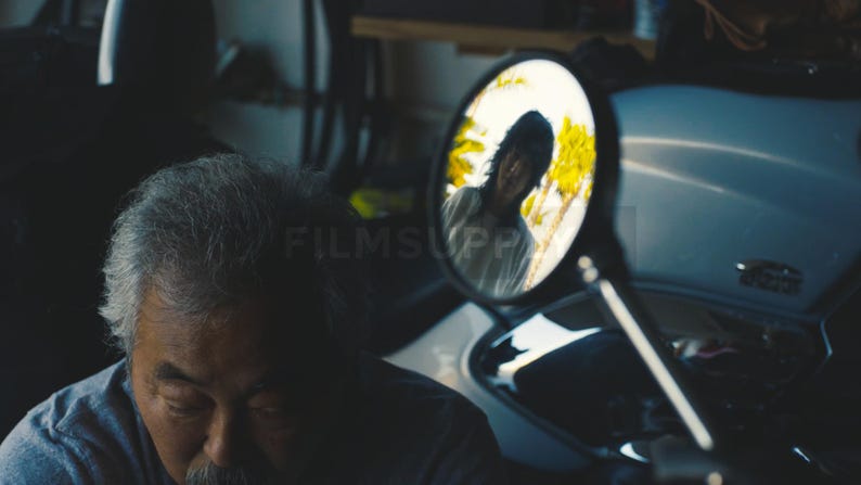May include: A person with grey hair and a moustache, looking down. A motorcycle mirror reflects a person in a white shirt. The scene is set in a garage with a motorcycle.