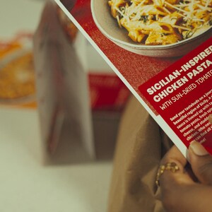 May include: A person holds a red and white food kit box with the text "SICILIAN-INSPIRED CHICKEN PASTA WITH SUN-DRIED TOMATO SAUCE." The box features a picture of pasta. A bag with food is in the background.