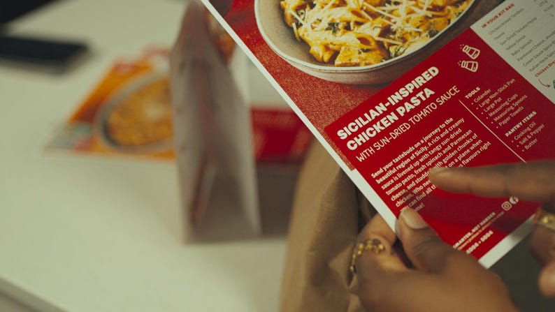 May include: A person holds a red and white food kit box with the text "SICILIAN-INSPIRED CHICKEN PASTA WITH SUN-DRIED TOMATO SAUCE." The box features a picture of pasta. A bag with food is in the background.