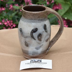 May include: A stoneware mug with a speckled brown rim and handle, featuring a white glazed body with dark brown brushstroke patterns. A white business card is placed in front, displaying the text LIBERTY STONEWARE and contact information.