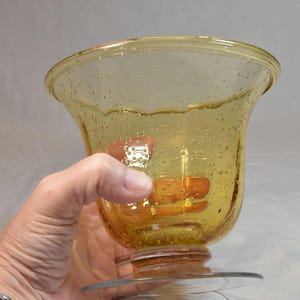 May include: A clear, yellow-tinted glass vase with a flared top and a round base. The vase has a bubble-like texture throughout the glass. The vase is held by a person's hand.