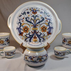 Peut inclure: Un service de table en c&eacute;ramique comprenant un grand plateau rond, deux tasses, deux petites tasses et un sucrier avec couvercle. L'ensemble est d&eacute;cor&eacute; d'un motif floral bleu, orange et jaune sur fond blanc. Le plateau a des poign&eacute;es.