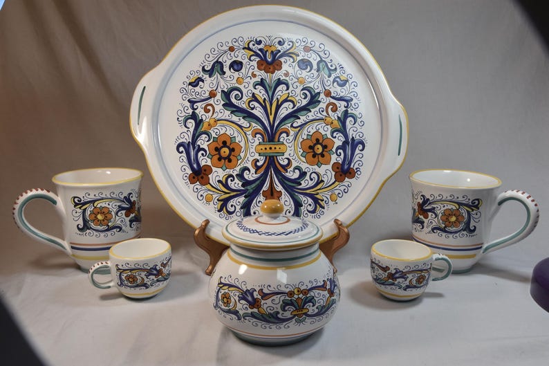 Peut inclure: Un service de table en c&eacute;ramique comprenant un grand plateau rond, deux tasses, deux petites tasses et un sucrier avec couvercle. L'ensemble est d&eacute;cor&eacute; d'un motif floral bleu, orange et jaune sur fond blanc. Le plateau a des poign&eacute;es.