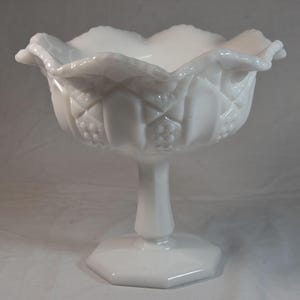 May include: A white, vintage-style pedestal bowl. The bowl has a scalloped rim and a textured, diamond and dot pattern on the sides. It sits on a stem and a hexagonal base. The piece is made of opaque white glass.