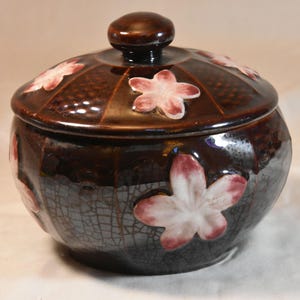 May include: A brown ceramic jar with a lid, adorned with pink and white flower designs. The jar has a crackle glaze finish and a small knob on top. The floral accents add a decorative touch to this storage container.