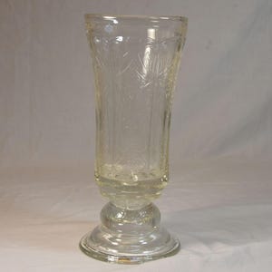 May include: A clear, vintage glass vase with a flared top and a pedestal base. The vase features an etched floral pattern and is approximately 10 inches tall. The base is round and has a decorative design.