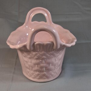 May include: A light pink ceramic basket featuring a woven texture on the exterior, a scalloped rim, and two arched handles. The vessel has a glossy finish and a rounded base, with a consistent pastel pink color throughout the entire structure.