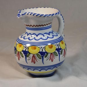 May include: White ceramic pitcher with a handle and spout, adorned with blue, yellow, and red floral patterns. The pitcher has a rounded body and a decorative blue trim around the rim and base.