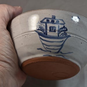 May include: A handmade ceramic bowl with a light gray exterior and a reddish-brown base. The bowl features a blue illustration of a boat with a house on top. The bowl is held in a hand.