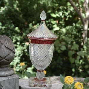May include: A clear glass lidded jar featuring a diamond-cut pattern across the body and lid, accented with a deep red band around the rim and base. The jar stands on a square glass pedestal base and is topped with a pointed, faceted glass finial.