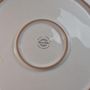 Peut inclure: Plat de service en c&eacute;ramique blanche avec un large bord l&eacute;g&egrave;rement sur&eacute;lev&eacute; et un centre plus petit et encastr&eacute;. Le bord et l'anneau central ont une bordure marron clair. Le plat porte le texte "Sur la table" et "Made in Italy".
