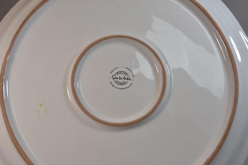 Peut inclure: Plat de service en c&eacute;ramique blanche avec un large bord l&eacute;g&egrave;rement sur&eacute;lev&eacute; et un centre plus petit et encastr&eacute;. Le bord et l'anneau central ont une bordure marron clair. Le plat porte le texte "Sur la table" et "Made in Italy".