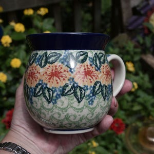 May include: A round ceramic mug with a dark blue rim and handle. The body features a repeating pattern of orange floral motifs, green leaves, and small blue dots, set against a cream-colored background with light green decorative scrollwork.