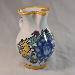May include: A white ceramic pitcher featuring a hand-painted floral design with a large blue flower and a smaller yellow flower. The pitcher has a yellow rim and a yellow base, with green leaves and purple accents surrounding the central floral motif.