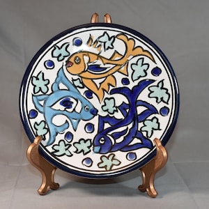 May include: A round ceramic plate featuring a hand-painted design of three fish in shades of orange, light blue, and dark blue. The background is white with small light green leaf-like shapes and dark blue dots, framed by a dark blue rim.