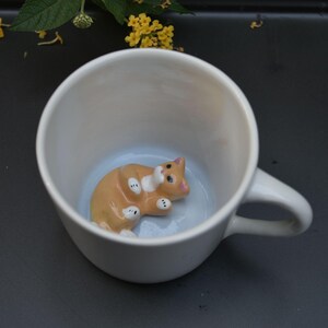 May include: A white ceramic mug featuring a small, three-dimensional orange and white cat figurine attached to the interior base. The cat is depicted in a lying position with its paws visible, and the mug has a smooth, glossy finish throughout.