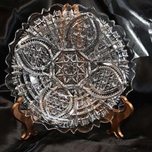 May include: A clear pressed glass plate featuring a central octagonal star pattern surrounded by four distinct petal-shaped sections. The plate has a scalloped edge and intricate geometric cross-hatch detailing throughout the entire surface.