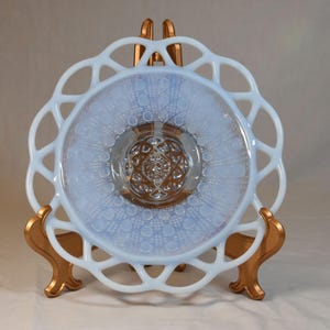 May include: A vintage, opalescent glass plate with a delicate, lacy edge. The plate features a light blue hue with a central, detailed design. It is displayed on a gold-colored metal stand, showcasing its intricate details and craftsmanship.