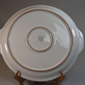 Peut inclure: Un plat de service en c&eacute;ramique blanche avec un design large et peu profond. Le bord et le cercle int&eacute;rieur ont une bordure marron. Le plat a deux poign&eacute;es et un centre plus petit et encastr&eacute;. Le fond du plat est visible.