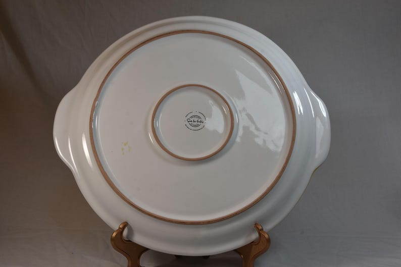 Peut inclure: Un plat de service en c&eacute;ramique blanche avec un design large et peu profond. Le bord et le cercle int&eacute;rieur ont une bordure marron. Le plat a deux poign&eacute;es et un centre plus petit et encastr&eacute;. Le fond du plat est visible.