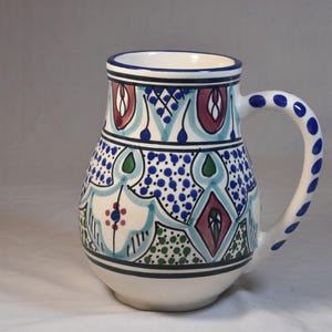 May include: A white ceramic pitcher with a handle, adorned with intricate hand-painted designs. The patterns include blue dots, teal and burgundy shapes, and green accents. The handle features blue dots.