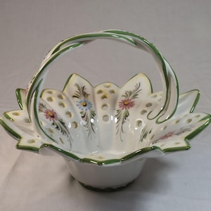 May include: A white ceramic basket with a green handle and rim. The basket has a scalloped edge and is decorated with small flowers in pink, blue, and yellow. The handle is twisted and has green accents.