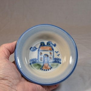 May include: A small, round ceramic bowl with a hand-painted house design. The bowl features a light blue rim and a cream-colored interior with a blue house, orange roof, and green fence. The bowl is held in a hand.
