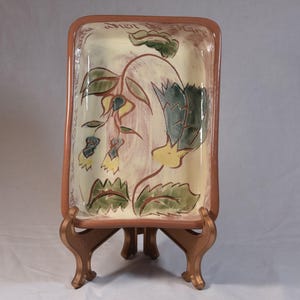 May include: Rectangular ceramic serving tray with a floral design in green, yellow, and blue. The tray has a light beige background with a brown border and is supported by a decorative bronze stand. The tray has text on the top border.