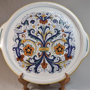 Peut inclure: Un plateau de service en c&eacute;ramique blanche avec un motif floral color&eacute;. Le plateau pr&eacute;sente un motif central de fleurs orange, de feuilles bleues et d'accents jaunes. Il a deux poign&eacute;es et un bord jaune.