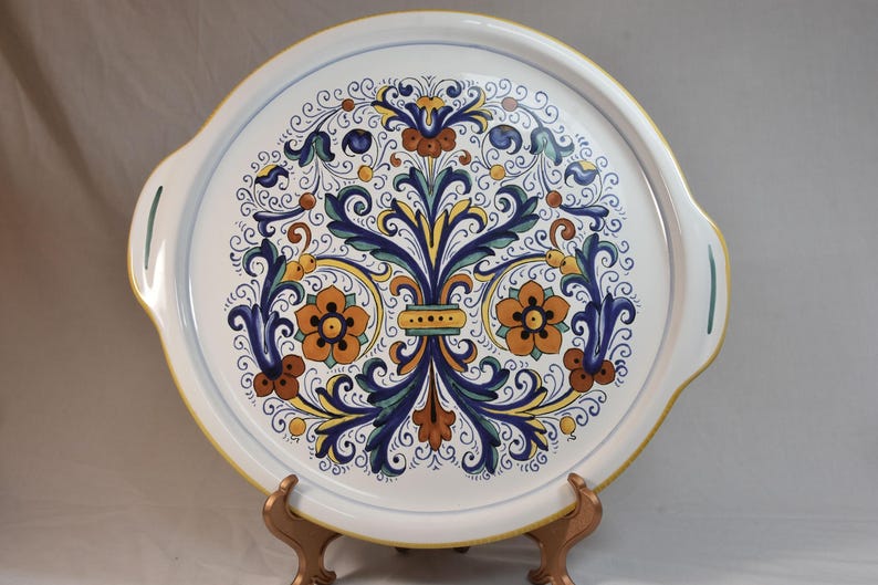 Peut inclure: Un plateau de service en c&eacute;ramique blanche avec un motif floral color&eacute;. Le plateau pr&eacute;sente un motif central de fleurs orange, de feuilles bleues et d'accents jaunes. Il a deux poign&eacute;es et un bord jaune.